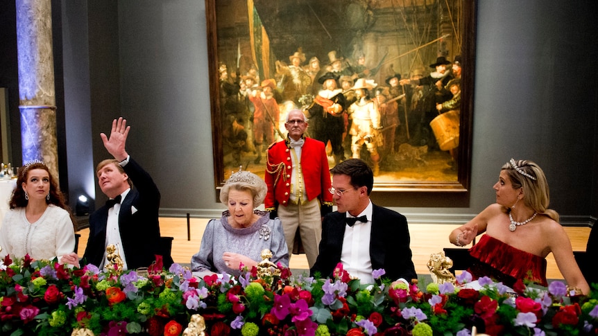 Four people in formal clothes sit at a long table decorated with flowers with a large oil painting in the background