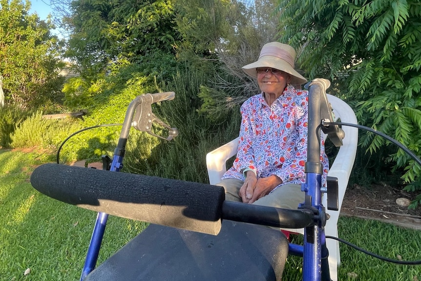 An elderly woman sitting in a chair in the garden, with her walker in front of her.