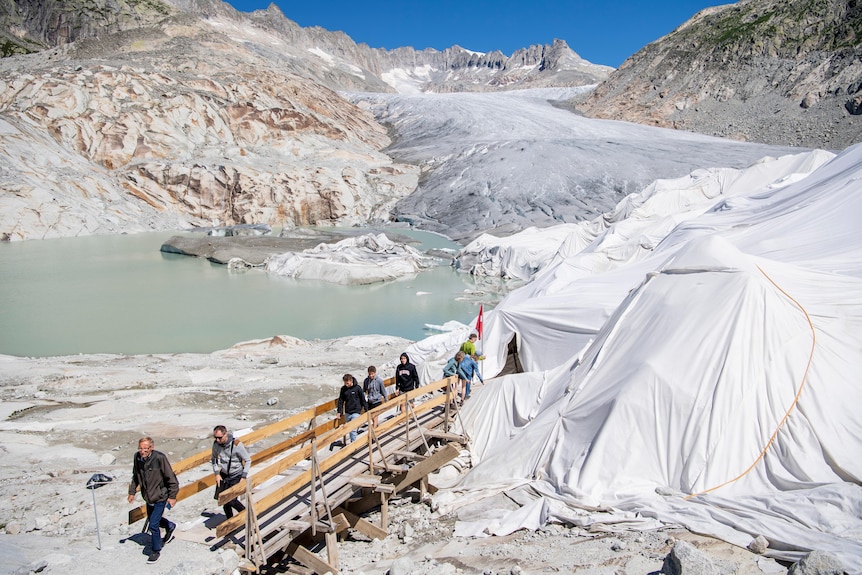 White blankets are draped over a portion of the alps as people walk across a wooden boardwalk to look inside. 