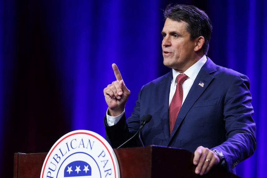 A middle-aged white man with dark hair and wearing a blue suit points as he speaks behind a lectern in front of a blue curtain.