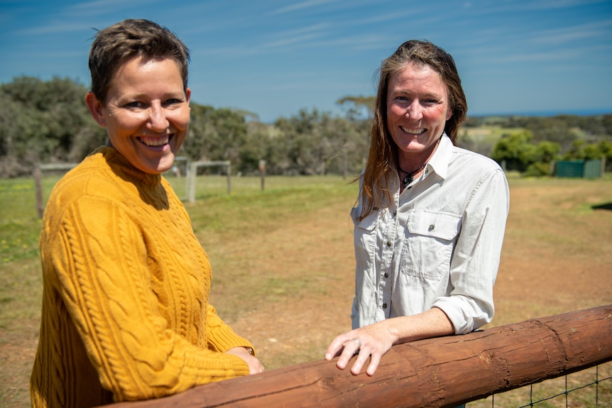 Two women standing outdoors in a rural setting, leaning on a fence and smiling.