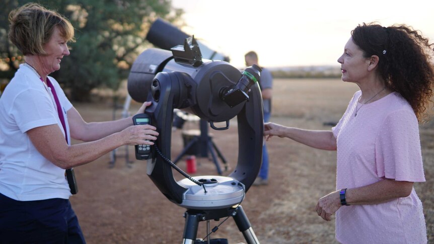 Carol Redford, left, stands with a remote in her hand as she moves a telescope next to a woman on her right.
