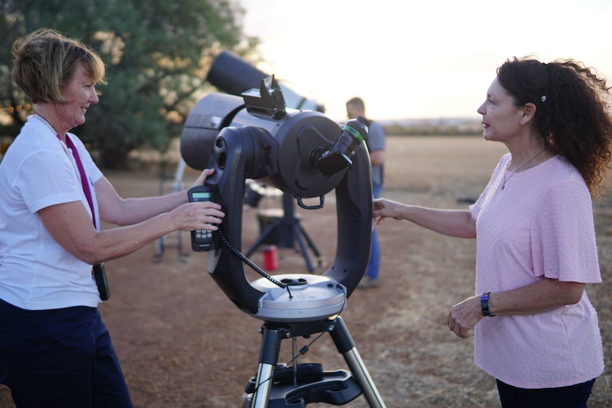Carol Redford, left, stands with a remote in her hand as she moves a telescope next to a woman on her right.
