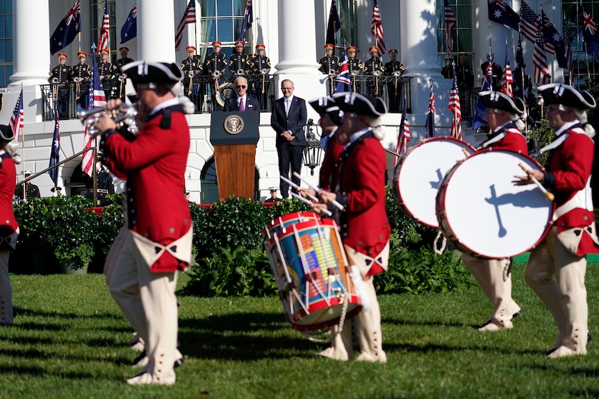 Band members with red jackets play drums and trumpets as Joe Biden and Anthony Albanese watch from a stage.
