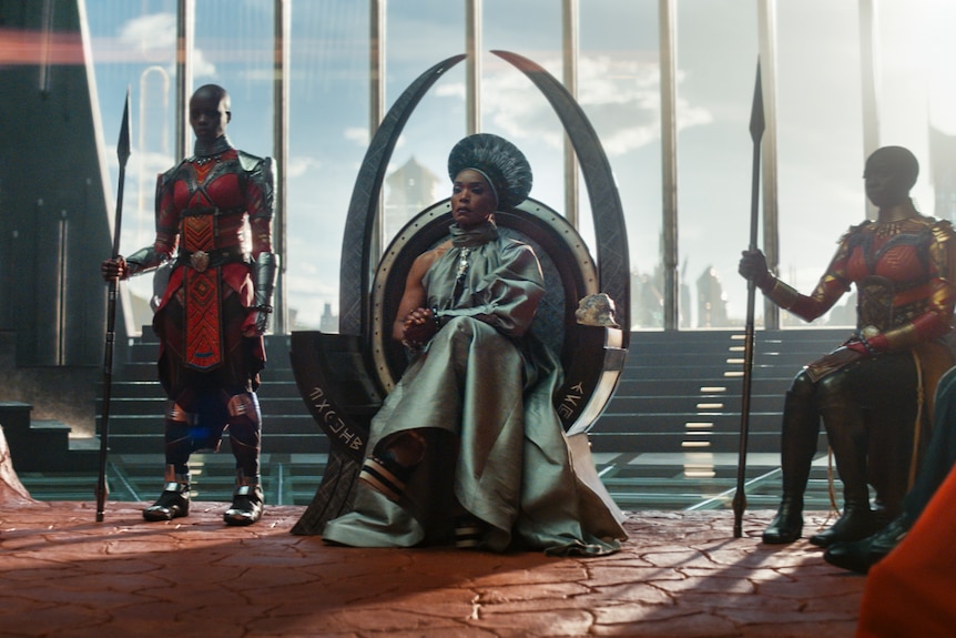 Black woman in futuristic-looking silver cape and large headdress sits in pincer-shaped chair with henchwomen either side.