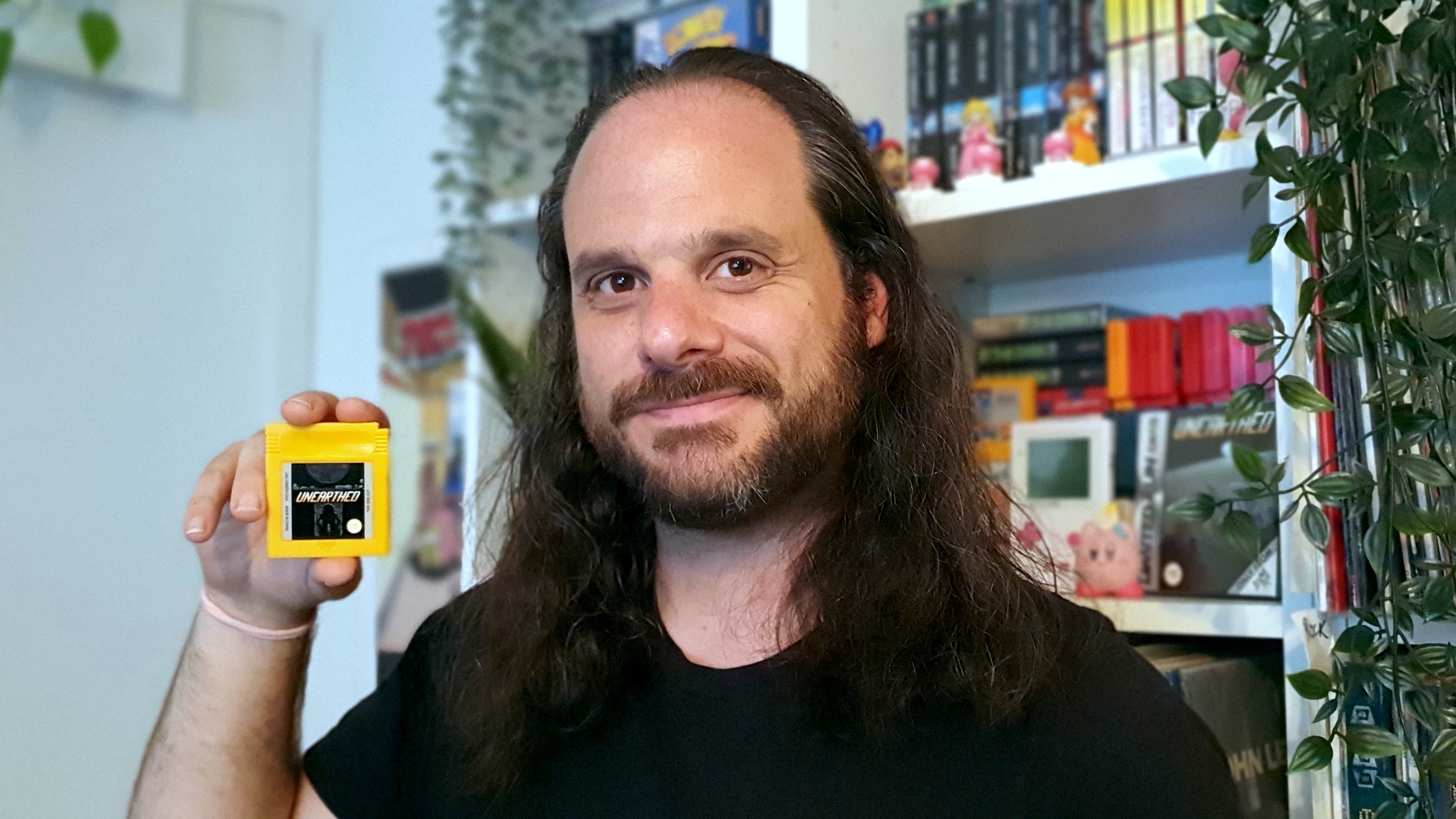 A man smiles towards the camera holding a yellow game boy cartridge.