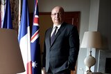 The French Ambassador wearing a suit stands beside a window next to Australian and French flags