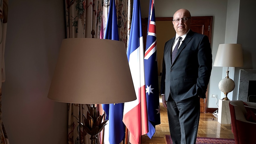 The French Ambassador wearing a suit stands beside a window next to Australian and French flags