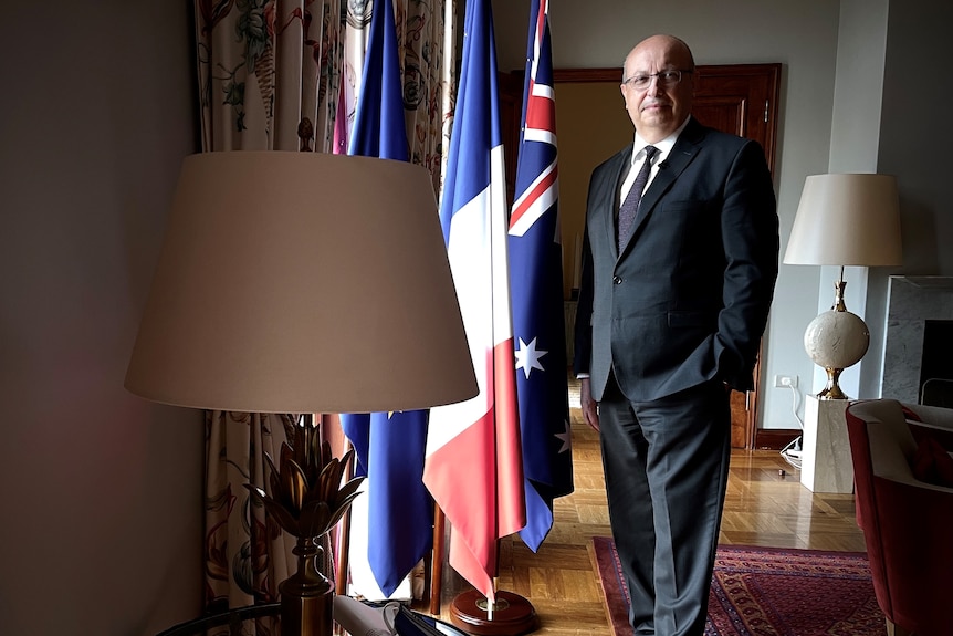 The French Ambassador wearing a suit stands beside a window next to Australian and French flags