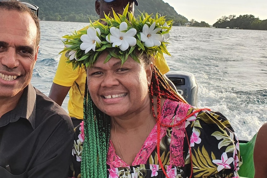 A woman with long briads wears a white and green floral headpiece while on a boat with a man at sunset