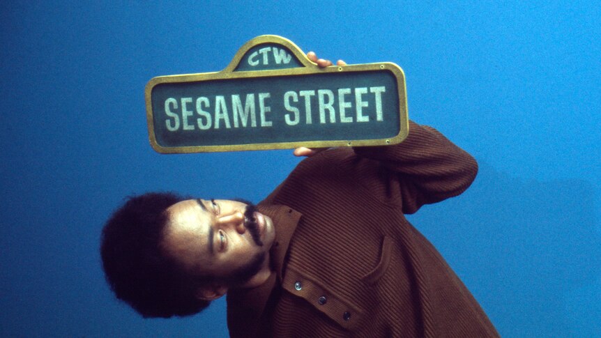 Young black man with moustache and short afro wears brown button-up shirt and hols street sign saying "Sesame Street"