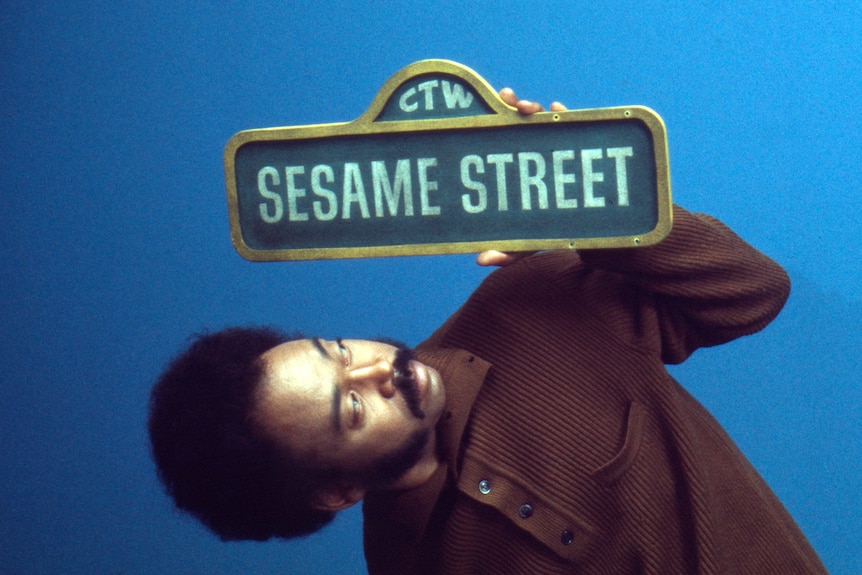 Young black man with moustache and short afro wears brown button-up shirt and hols street sign saying "Sesame Street"