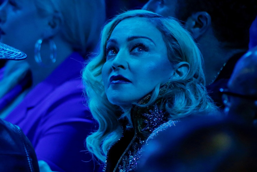 Madonna, sitting during an awards show, looks in the distance as blue light dims the hall.