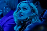 Madonna, sitting during an awards show, looks in the distance as blue light dims the hall.