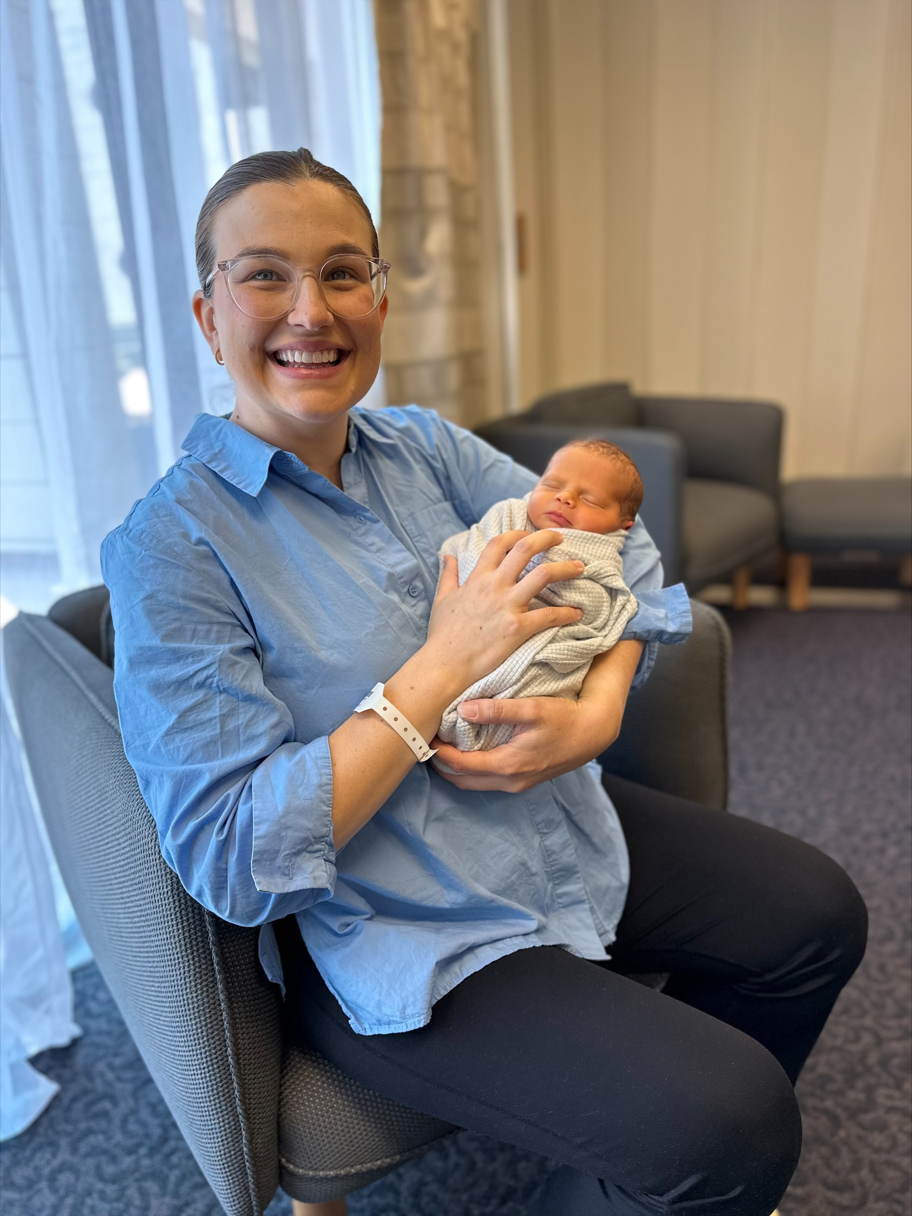 A woman sits smiling, holding a newborn baby.