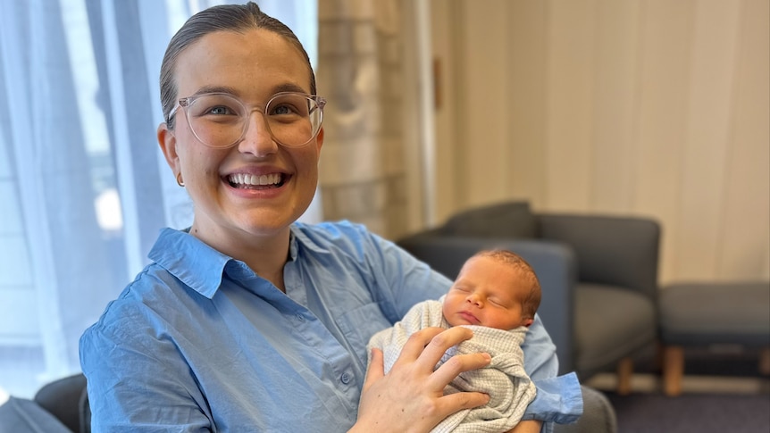 A woman sits smiling, holding a newborn baby.