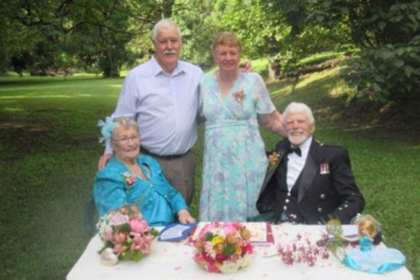 woman in blue and man in black jacket sit at table with flowers and man in light shirt and woman dress stand with them