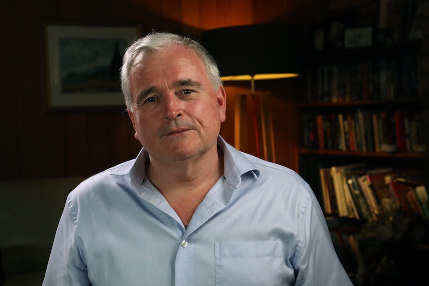 A man sitting in a room with a bookshelf, looks at the camera.