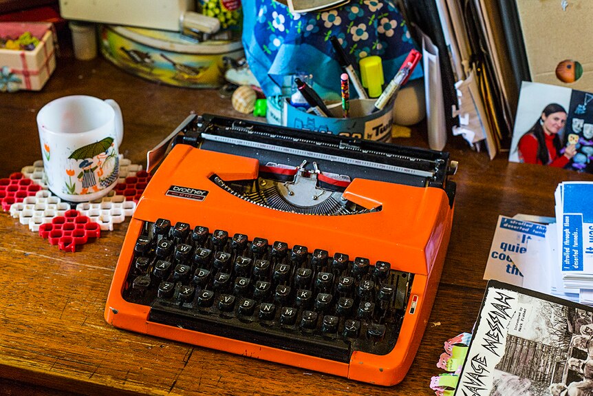 A bright orange, 1960s style typewriter sits on a desk surrounded by knickknacks.