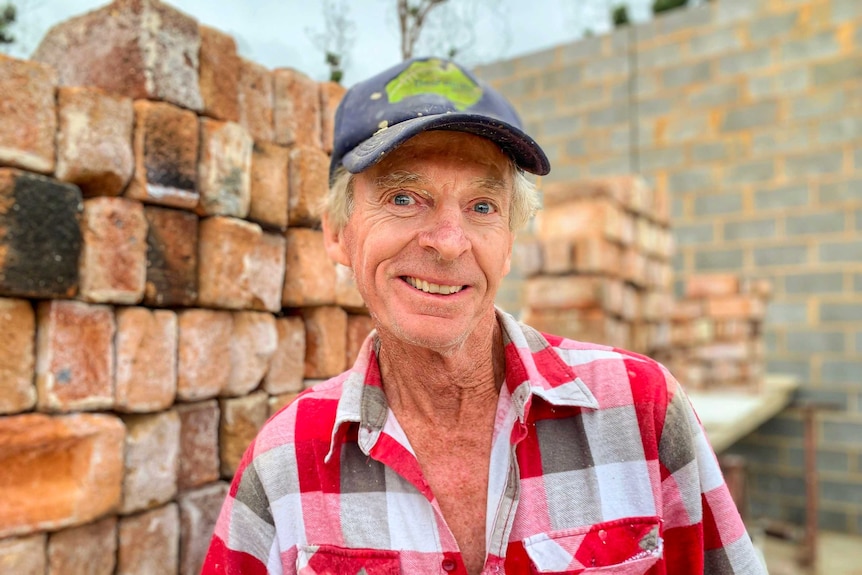 A man in a red flannel shirt stands in front of a brick wall and piles of bricks.