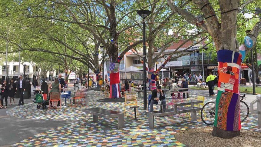 A plaza with colourful chairs, trees wrapped in colourful wool and painted pavement.