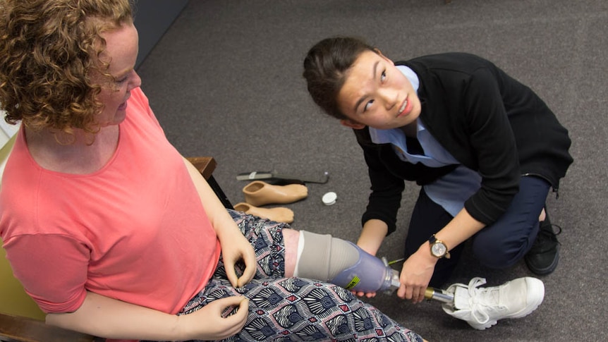 A woman with prosthetic arms and legs sits in a chair. A young woman crouches beside, holding the sitting woman's left leg.