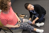 A woman with prosthetic arms and legs sits in a chair. A young woman crouches beside, holding the sitting woman's left leg.