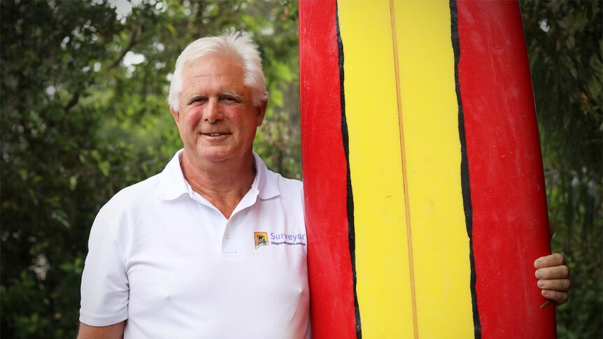 Ken Holloway holds a red and yellow surf board upright while smiling at the camera.