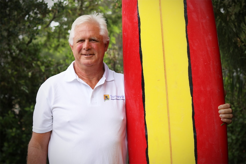 Ken Holloway holds a red and yellow surf board upright while smiling at the camera.