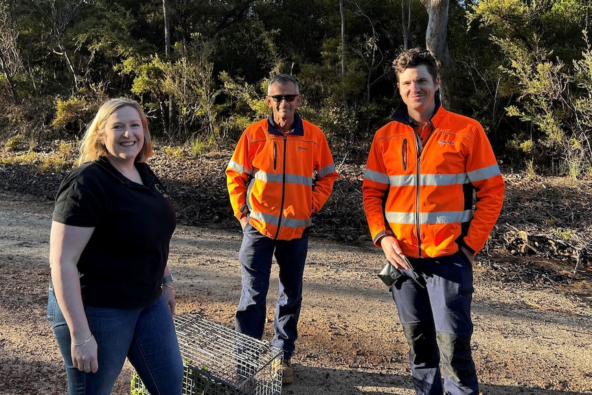 A woman and two men smile at the camera while standing in bushland.