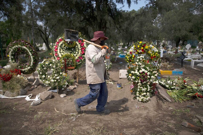A man wearing a face mask carries a shovel over his right shoulder as he walks past a row of decorated graves.