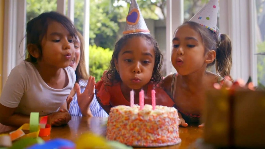 Three girls, two wearing party hats, blowing out the candles on a cake