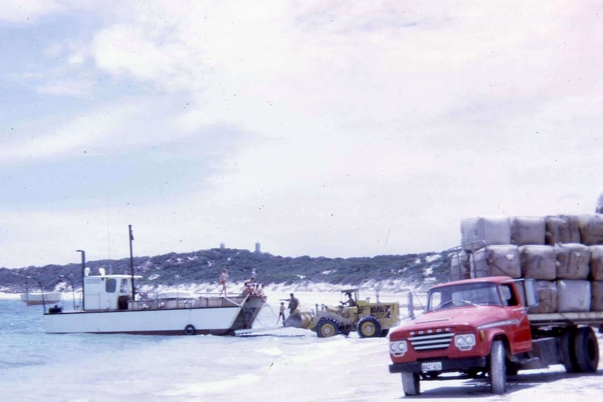 An old photograph of men unloading wool on a barge with a tractor on the beach receiving the stock.