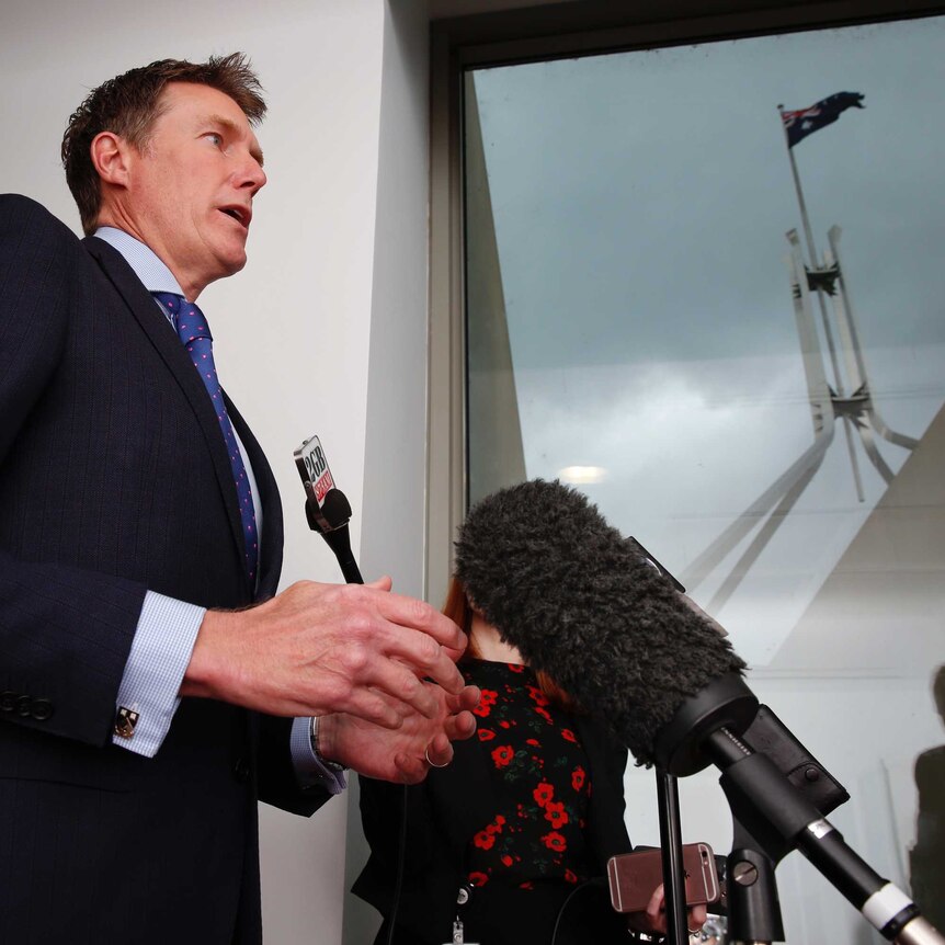 Christian Porter speaks into microphones. Behind him, through a window, the Parliament House flag is seen against a stormy sky.