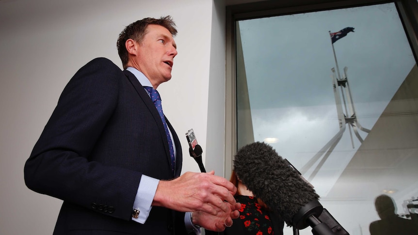 Christian Porter speaks into microphones. Behind him, through a window, the Parliament House flag is seen against a stormy sky.
