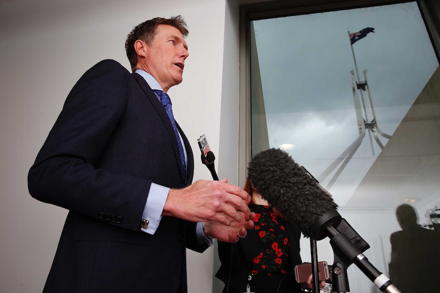 Christian Porter speaks into microphones. Behind him, through a window, the Parliament House flag is seen against a stormy sky.