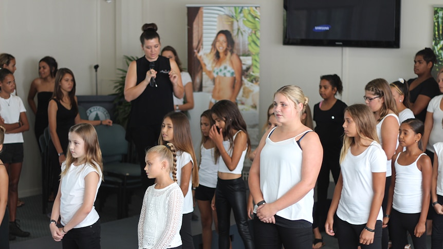 Around twenty young Aboriginal girls stand, learning how to pose like a model.