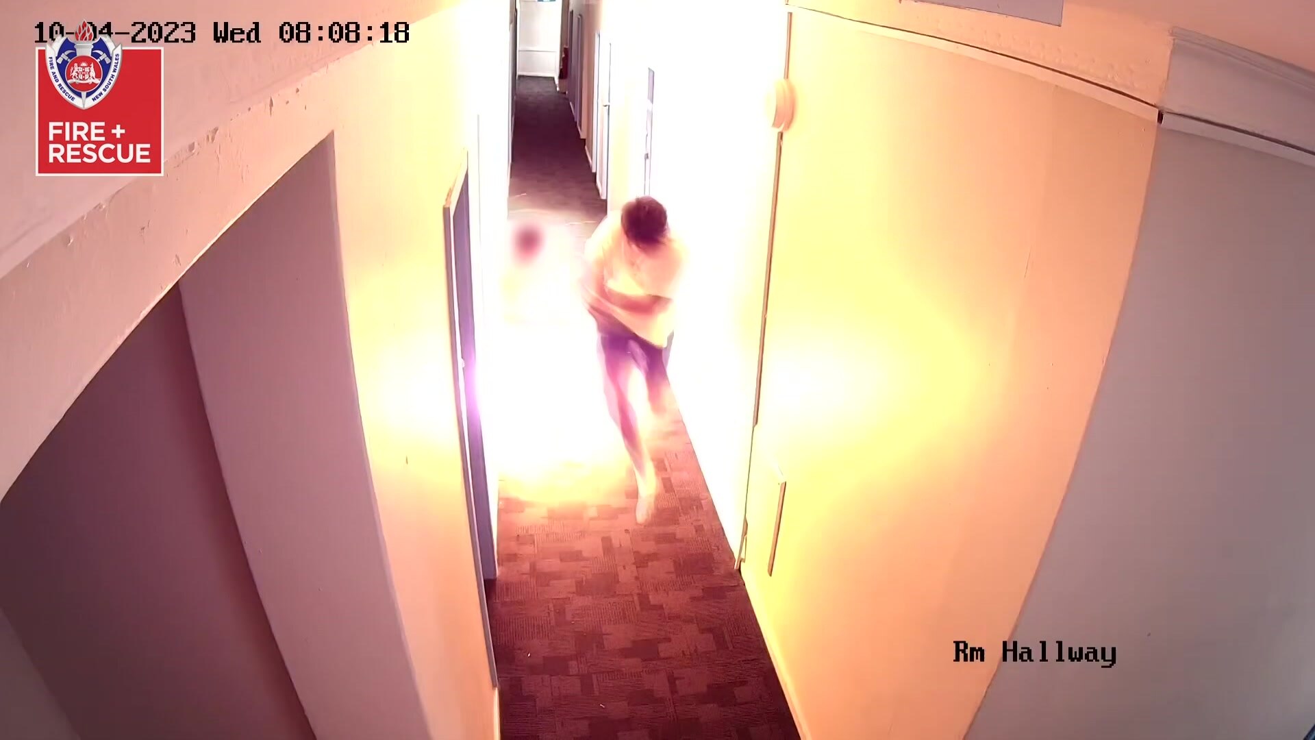 A man standing in a narow hallway shields himself as flames explode out of a doorway 