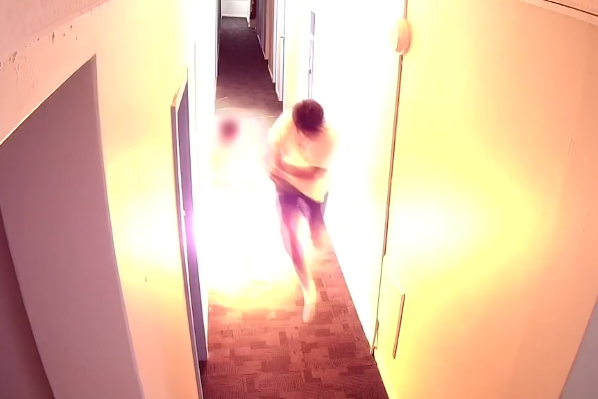 A man standing in a narow hallway shields himself as flames explode out of a doorway.