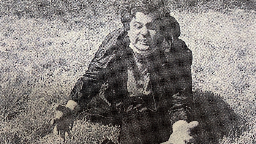 A black and white image of a man squatting down in a field.