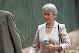 A woman with short grey hair walks while wearing a pale blazer and carrying a water bottle.