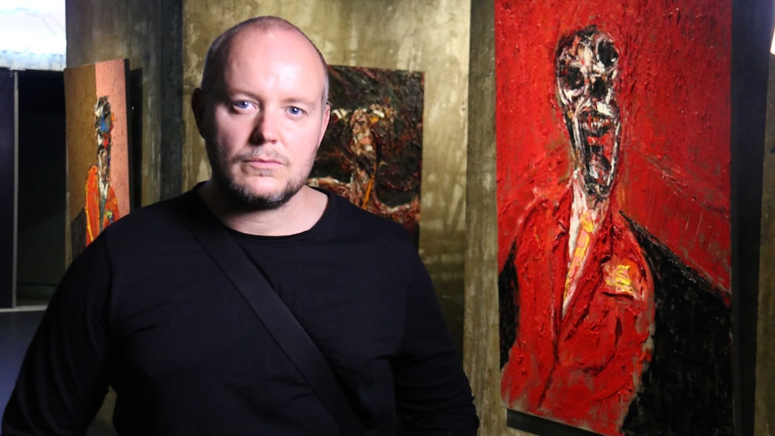 Artist Lincoln Townley stands in front of his artworks hanging on concrete wall at a Brisbane exhibition.