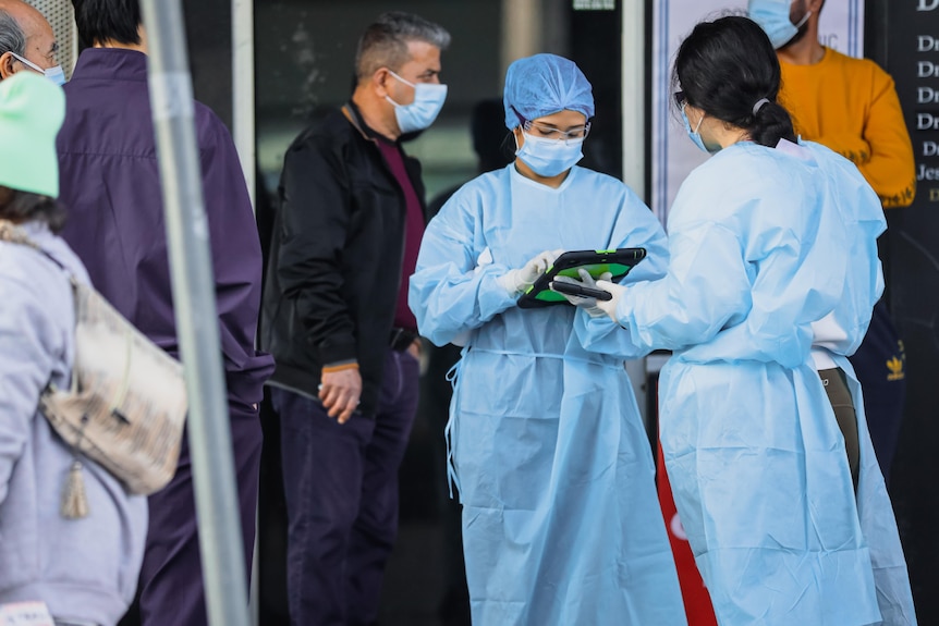 Two women wearing protective gowns and face masks check a clipboard while a man walks past in the background.