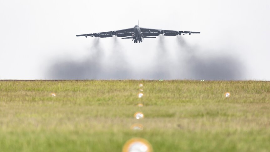 A large jet takes off from a runway, plumes of smoke trailing beneath it.