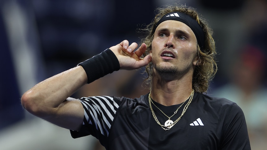 A German tennis player looks off to the side of the court as he cups his hand to his ear as if to hear what is being said.