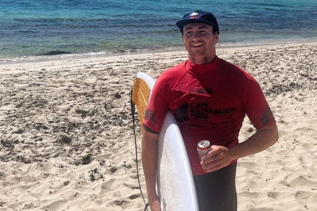 Max holds a surfboard on a beach wearing a red rash vest