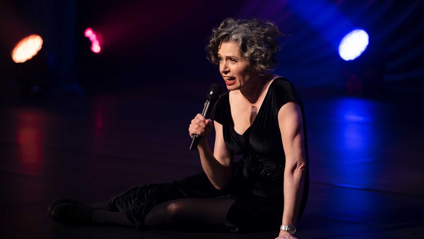 The comedian Judith Lucy sitting down on stage, microphone in hand