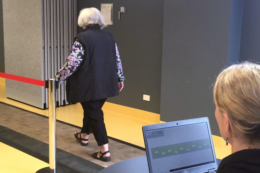 Elderly woman walks away from camera as researcher looks on while using laptop.