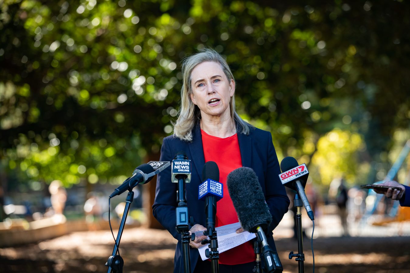 Simon McGurk pictured in front of trees speaking into microphones wearing a blazer and red top