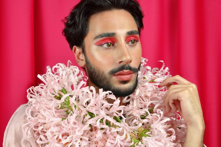 A man sits with a pink flower necklace with red eye shadow.
